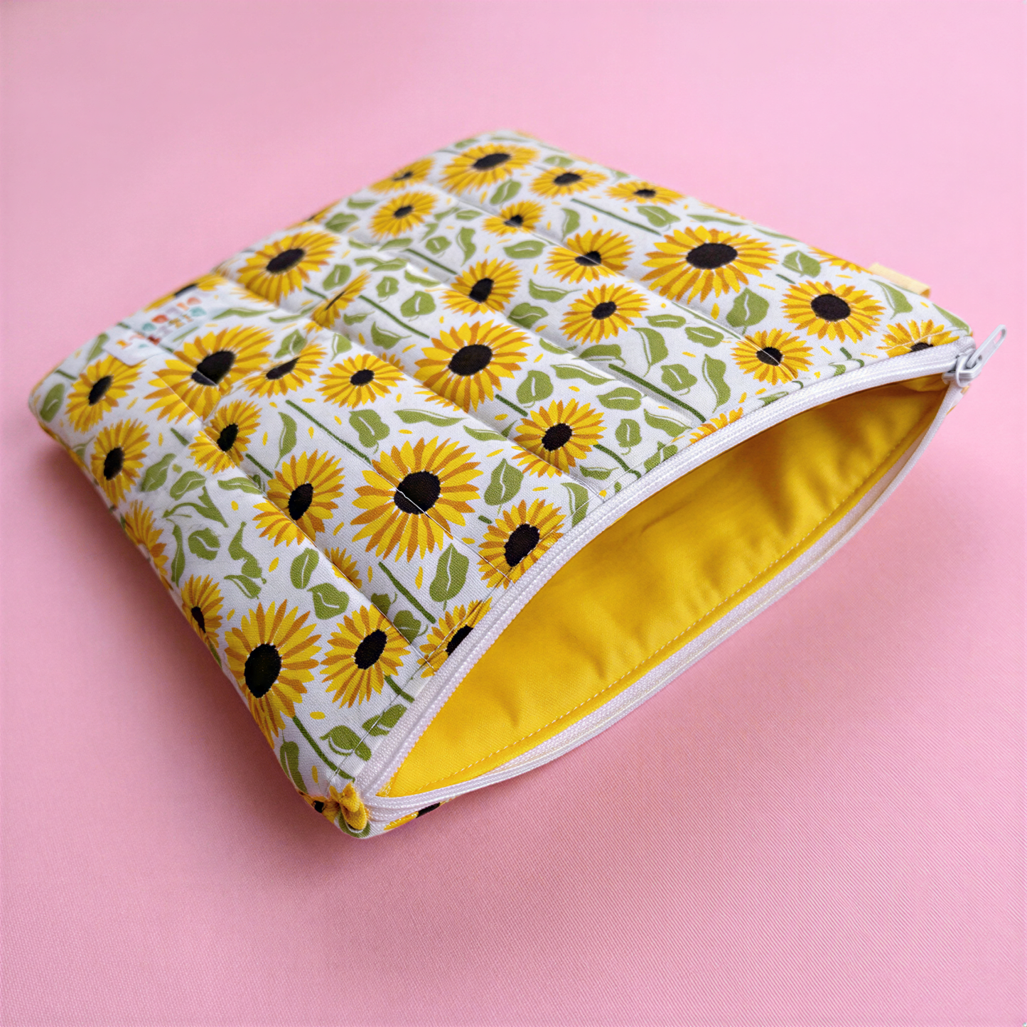 A white and yellow pouch with a sunflower pattern is shown against a pink background.