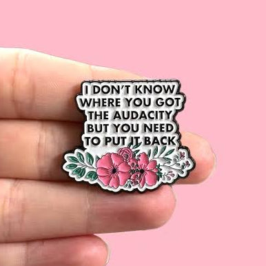 A hand holding a small enamel pin with the text 'I DON'T KNOW WHERE YOU GOT THE AUDACITY BUT YOU NEED TO PUT IT BACK' above pink flowers and green leaves, set against a solid pink background.