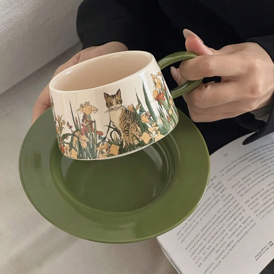 Person holding a ceramic mug with floral and cat design on a saucer